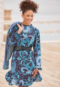 Langärmliges, mit Paisleymuster in Marineblau und Türkis versehenes Kleid mit einem voluminösen Saum und einem schwarzen Gürtel. Jeansjacke über eine Schulter drapiert.
