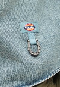 Υφασμα τζιν με ένα ανοιχτό μπλε κουμπί με το λογότυπο "Dickies" και μια ασημένια διακοσμητική πέταλο από κάτω, εμφανίζοντας ραμμένα άκρα και υφή.