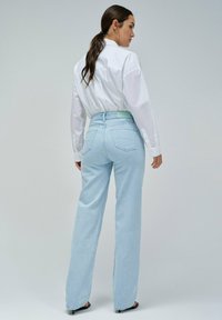 Jeans de pernera ancha en azul claro con cintura alta, que cuentan con bolsillos traseros tipo parche y costuras visibles. Combinados con una camisa blanca de manga larga.