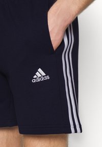 Jūras zila sporta šorti ar baltu trīsrindas dizainu, kabatām un izšūtu Adidas logo apakšējā kreisajā kājā. Mīksta auduma tekstūra.