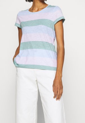 Kvinde iført pastelrosa, grøn og blå stribet kortærmet t-shirt med hvide bukser, hånd i lommen, mod en ensfarvet baggrund.