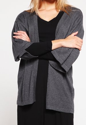 Cardigan grigio in stile kimono con maniche ampie e apertura sul davanti. Il tessuto testurizzato si adagia liberamente su un indumento nero sottostante.