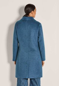 Vrouw met donker haar in een lage paardenstaart, gekleed in een lange blauwe jas en een patroon op blauwe broek, staande voor een effen lichtkleurige muur.