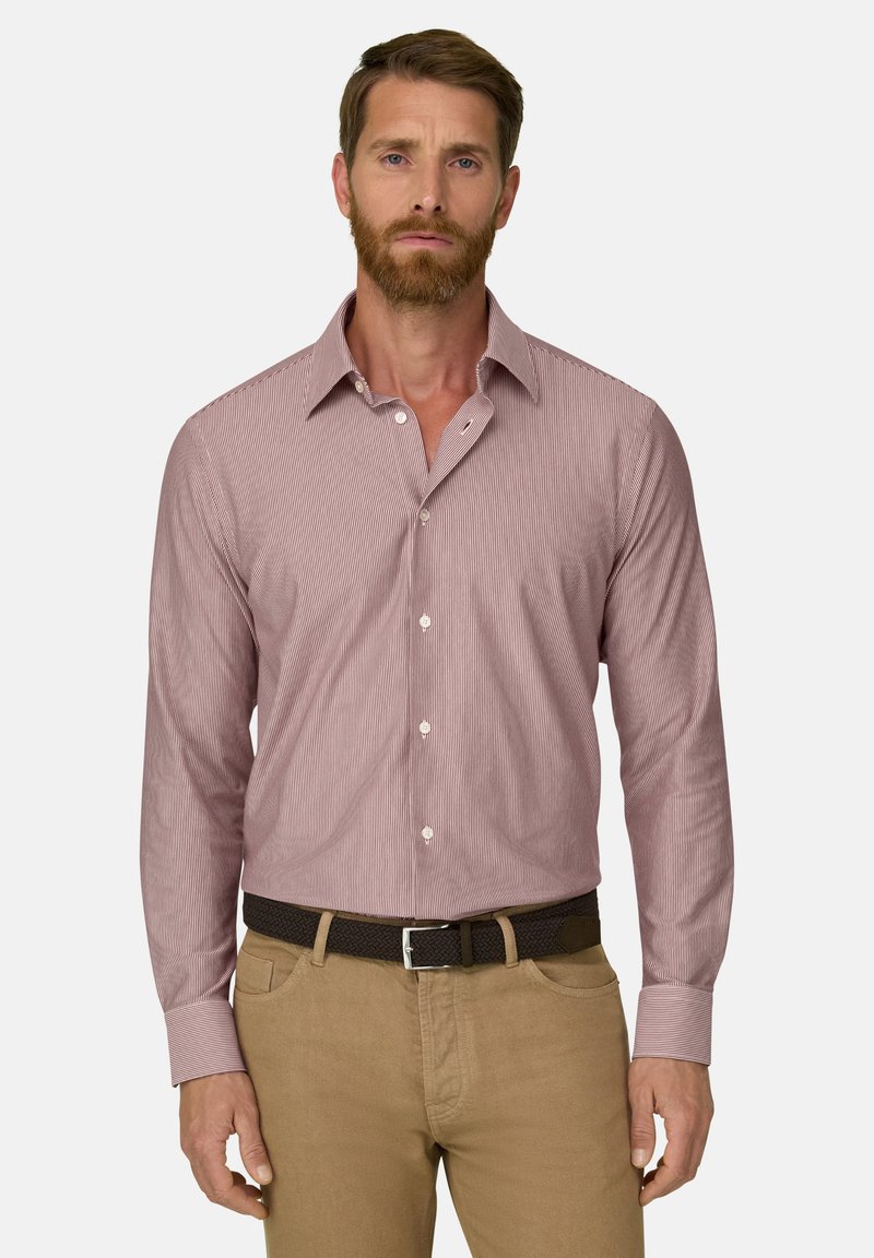 Homme aux cheveux bruns courts et portant une barbe, vêtu d'une chemise à rayures rouges claires boutonnée et d'un pantalon beige avec une ceinture noire, debout devant un fond uni.