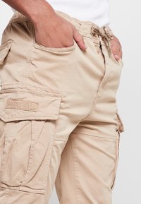 Pantalons cargo beige en coton doux, dotés d'une taille à cordon de serrage, de multiples poches et d'un patch logo visible sur le côté.