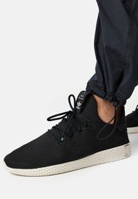 Zapatillas de malla negras con superficie texturizada, suela blanca en contraste, diseño con cordones y detalle del logo en la lengua.