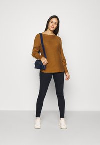 Brun strikket genser med vid halslinje, lange ermer, kombinert med mørke skinny jeans og hvite joggesko, holder en marineblå crossbody-veske.