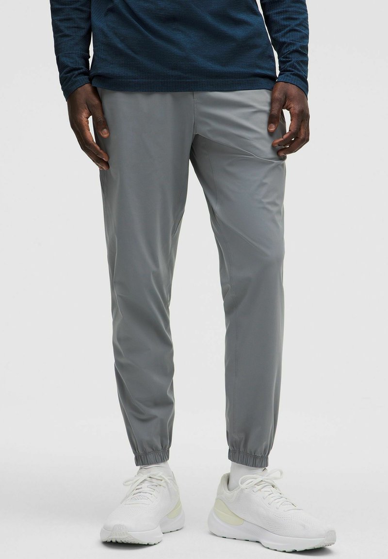 Pantaloni jogger leggeri, grigi, con polsini elasticizzati, tessuto traspirante e tasche laterali, abbinati a scarpe sportive bianche.