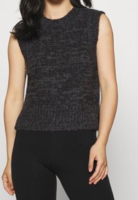 Donna che indossa un top senza maniche in maglia grigia scura e morbida abbinato a pantaloni neri aderenti, in piedi con le braccia rilassate ai lati.
