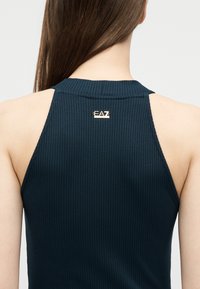 Γυναίκα με μακριά καστανά μαλλιά που φοράει αμάνικη μπλούζα σε σκούρο μπλε με ριμπ ύφανση, με ένα μικρό χρυσό λογότυπο EA7 στο πάνω μέρος της πλάτης.