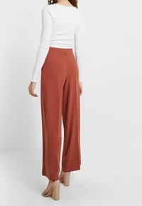 Femme portant un pantalon large taille haute couleur rouille, un haut ajusté à manches longues blanc et des sandales à talons blocs beige, se tenant devant un fond blanc.