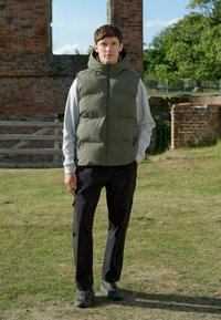 Olivegrüner Steppweste mit hohem Kragen, kombiniert mit einem hellgrauen Langarmshirt und schwarzen Cargo-Hosen. Strukturierten Stoff und sichtbare Reißverschlüsse.