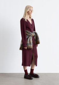 Langes bordeauxfarbenes Strickkleid mit tiefem V-Ausschnitt, vorderen Knöpfen und einem mit Gürtel versehenen Kunstfellbesatz. Es hat lange Ärmel und einen Oberschenkel-hohen Schlitz.