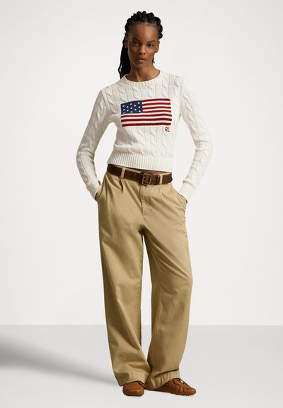 Oprijet krem pulover z rdeče-modrim ameriškim zastavnim vzorcem, kombiniran s širokimi hlačami v barvi kaki in rjavimi mokasinskimi čevlji.