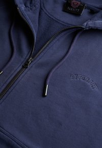 Granatowa bluza z kapturem zapinana na zamek, z czarnym zamkiem, sznurkami i haftowanym logo "SUPERDRY" na piersi, z metką "Varsity" wewnątrz kołnierza.