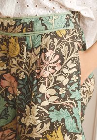 Pantalons à motifs floraux en tissu sombre, avec une taille verte, des poches latérales et des motifs floraux complexes en rose, jaune et bleu.