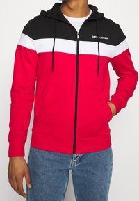 Červená mikina na zip s černou kapucí a bílým pruhovaným vzorem. Má šňůrky na stažení, přední kapsy a žebrované manžety.