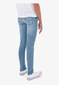 Persona con pantalones ajustados de mezclilla azul claro y zapatillas blancas, de pie frente a un fondo blanco liso, vista desde atrás.