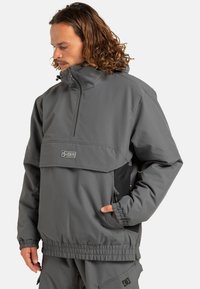 Szary sweter z wysokim kołnierzem, zamkiem błyskawicznym, elastycznymi mankietami i dołem. Posiada przednią kieszeń oraz detal z logo na klatce piersiowej.