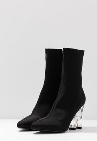 Une paire de bottines noires avec des pointes en amande et des talons blocs transparents sur un fond blanc et gris clair.