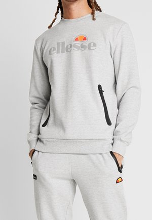 Persona vestida con sudadera ellesse gris claro y pantalones jogger a juego con bolsillos con cremallera negra y logo de la marca en el pecho y el muslo.