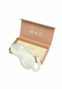 Siden ögonmask i ljus grädde med elastiskt band, förpackad i en persikofärgad låda med guldiga ikoner och präglad text.