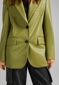 Olivově zelená faux kůže blejzr s upraveným střihem, dvěma p�ředními kapsami a černými knoflíky, kombinovaný s černými cargo kalhotami.