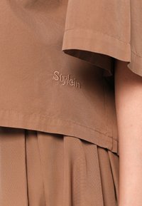 Brown short-sleeve top made of textured fabric, featuring a layered design and subtle "Stylein" embroidery on the front.