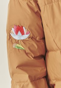 Brūns pūkains jaka ar krāsainu izšūtu lotosa ziedu uz kreisā roka, izceļot sarkanas, baltas, rozā un zaļas diegus.