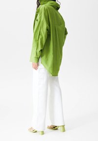 Chemise verte surdimensionnée avec une finition texturée, des manches longues et un ourlet haut-bas, associée à un pantalon large blanc et des sandales à talons blocs vertes.