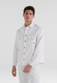 Chaqueta de mezclilla blanca con puños ajustables, dos bolsillos en el pecho y costuras en contraste. Presenta botones metálicos y un acento de logo.