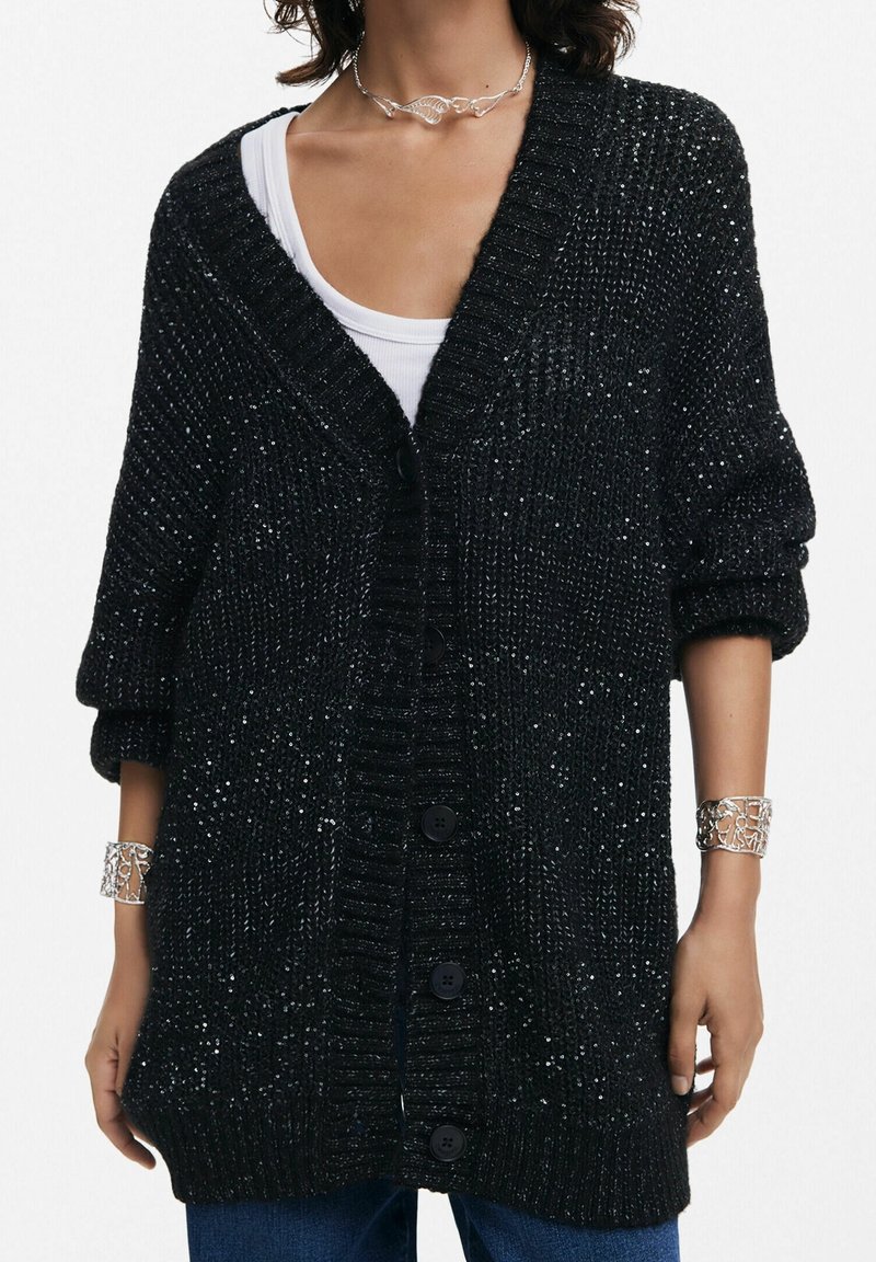 Femme portant un cardigan en maille épaisse noire avec de gros boutons, un top blanc en dessous, un collier feuille argenté et des bracelets manchettes assortis en argent.