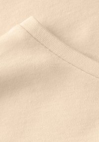 Beige látka s hladkou strukturou; obsahuje žebrovaný límec a mírný šikmý šev. Detailní pohled zdůrazňuje měkkou pletenou kvalitu.