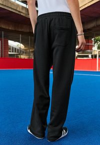 Schwarze Sweatpants mit entspanntem Schnitt, elastischem Bund und einer einzelnen Gesäßtasche, kombiniert mit schwarz-weißen Schuhen auf einer blauen Oberfläche.