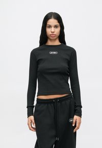 Femme avec de longs cheveux noirs portant un crop top noir à manches longues en côtes et un pantalon noir à cordon de serrage, tous deux arborant un petit logo blanc "jordan".