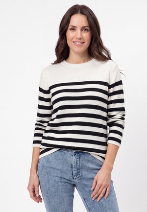 Kvinde med langt brunt hår iført en sort og hvid vandretstribet sweater og lyseblå jeans, smilende med en neutral hvid baggrund.