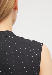 Blusa nera senza maniche con un motivo a pois bianchi. Presenta una texture liscia e un sottile design del colletto sul retro.