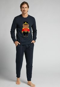 Navy souprava pyžama obsahuje dlouhý rukáv s grafickým motivem plyšového medvídka v červené a zlaté, doplněná o odpovídající kalhoty se stahovacími manžetami na kotnících.