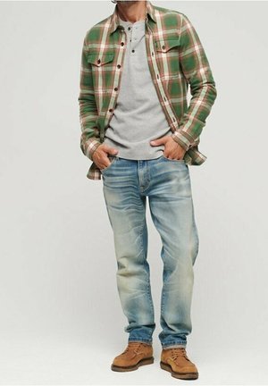 Homme portant une chemise à carreaux verte, un henley gris, un jean bleu clair et des bottes à lacets couleur sable, debout les mains dans les poches.