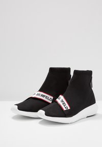 Černé pletené kotníkové boty s bílou gumovou podrážkou a červeným a bílým popruhem se značkou. Mají přiléhavý design ponožky a minimalistické detaily.