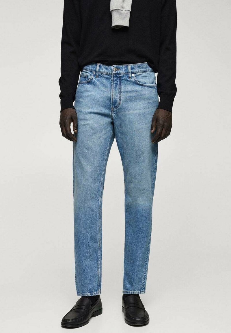 Personne portant un jean droit bleu clair, des mocassins noirs et un haut noir à manches longues avec des manches grises tombant sur les épaules.