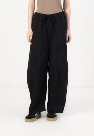 Persoon die losse zwarte cargobroeken met een trekkoord in de taille en zwarte sneakers met witte strepen en rubberen zolen draagt, staand tegen een witte achtergrond.