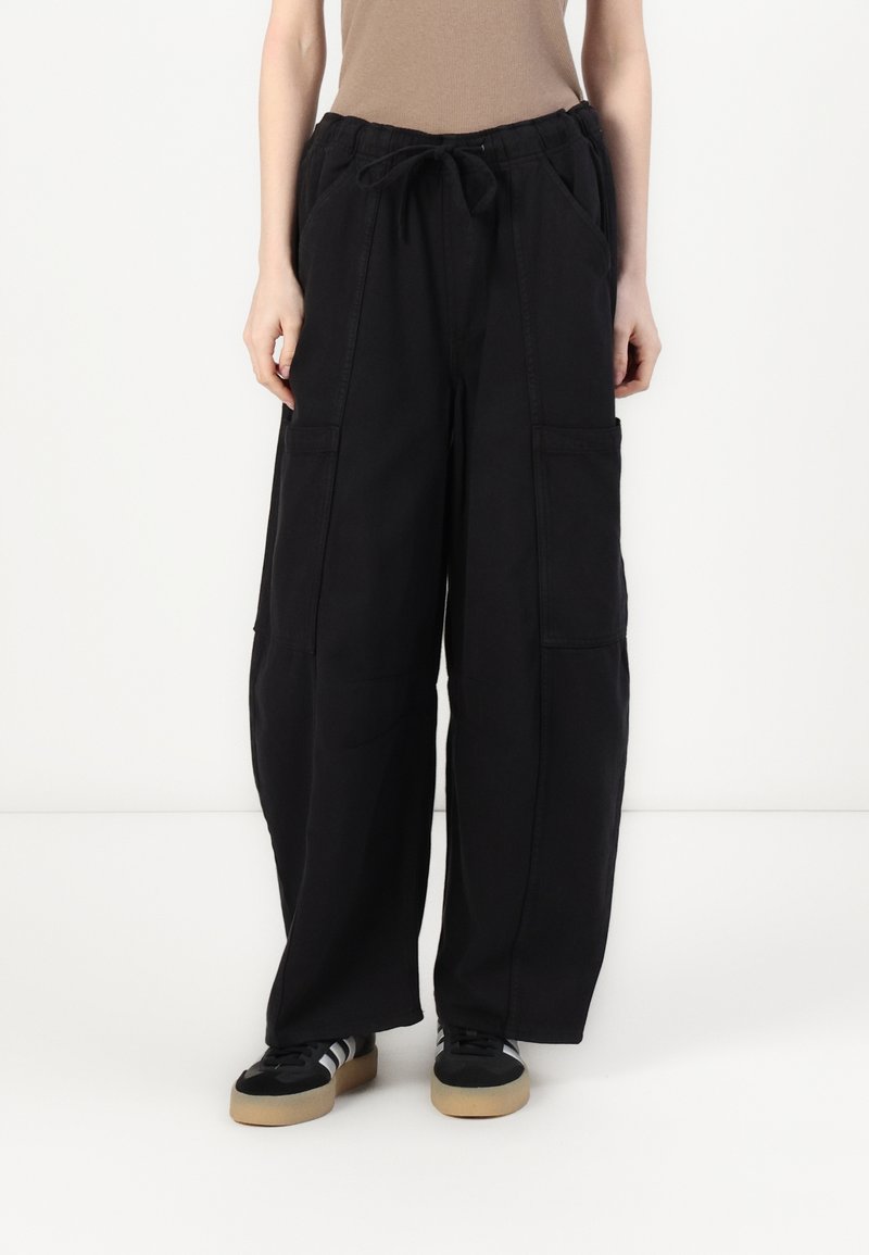 Personne portant un pantalon cargo noir ample avec une taille à cordon et des baskets noires avec des bandes blanches et des semelles en gomme, debout devant un fond blanc.