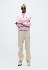 Άτομο στέκεται με σταυρωμένα τα χέρια, φορώντας φούτερ με φερμουάρ στο λαιμό σε ανοιχτό ροζ χρώμα, μπεζ παντελόνι καρότσα, μπεζ αθλητικά παπούτσια και μεγάλα γυαλιά ηλίου με αντανακλαστικούς φακούς.