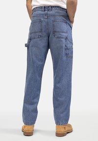 Homme portant un jean de charpentier bleu en denim avec plusieurs poches arrière et latérales, des bottines de travail marron clair et une chemise blanche, vu de dos.