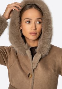 Brązowy zamszowy płaszcz z kapturem z sztucznego futra, z zapięciem na guziki i teksturowanymi akcentami. Kołnierz i kaptur mają miękki, puszysty wygląd.