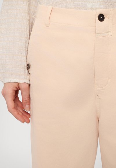 Pantaloni de culoare piersică deschisă, cu o textură netedă din bumbac. Prezintă o închidere cu nasture, un buzunar lateral cu clapetă metalică și detalii de cusătură subtile.