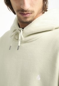 Lichtgroene hoodie van zacht materiaal, met een trekkoordcapuchon en een klein wit logo op de borst, met zilverkleurige punten aan de uiteinden van het trekkoord.