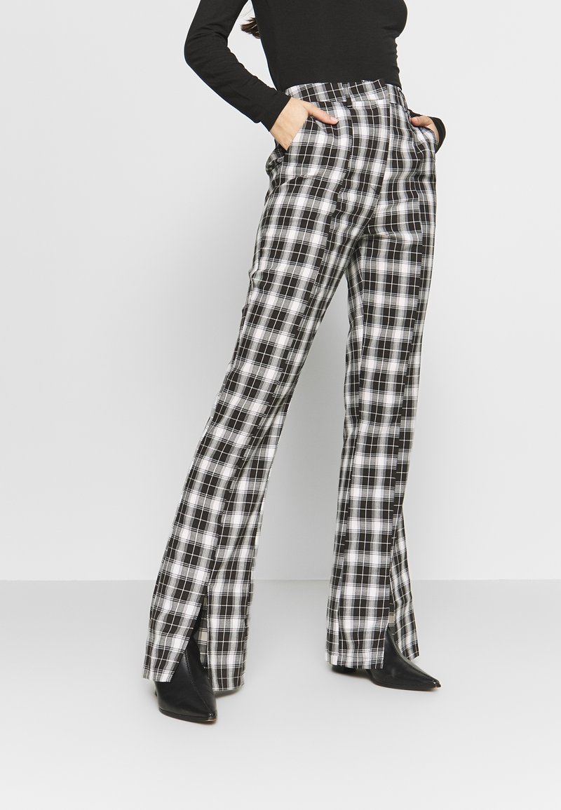 Personne portant un pantalon à carreaux noir et blanc taille haute avec des fentes latérales et des bottes noires à bouts pointus, mains dans les poches, debout devant un fond uni.