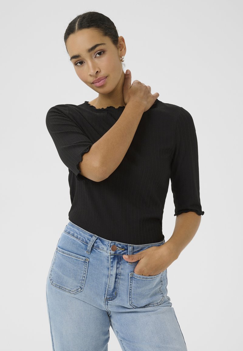 Sort ribbet top med korte flæseærmer, parret med lyseblå højtaljede jeans; modellen står med hånd på nakken. Simpelt design, klassisk pasform.